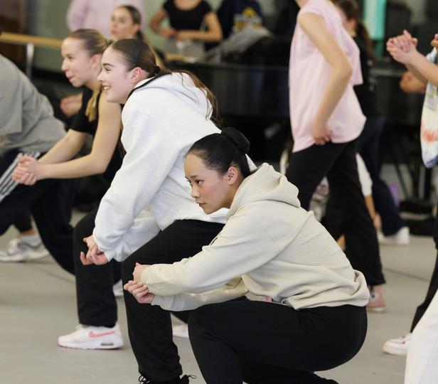 A group of young dancers in a dance class. The dancers, wearing casual dance attire including hoodies and sweatpants, are engaged in a dynamic pose, some crouching low to the ground. They appear focused and enthusiastic, with some smiling and others deeply concentrating on their movements.